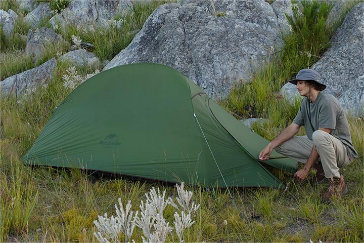 A scene of a man sitting in front of a tent. The tent is green and the man is sitting on the ground. He is checking the entrance of the tent.