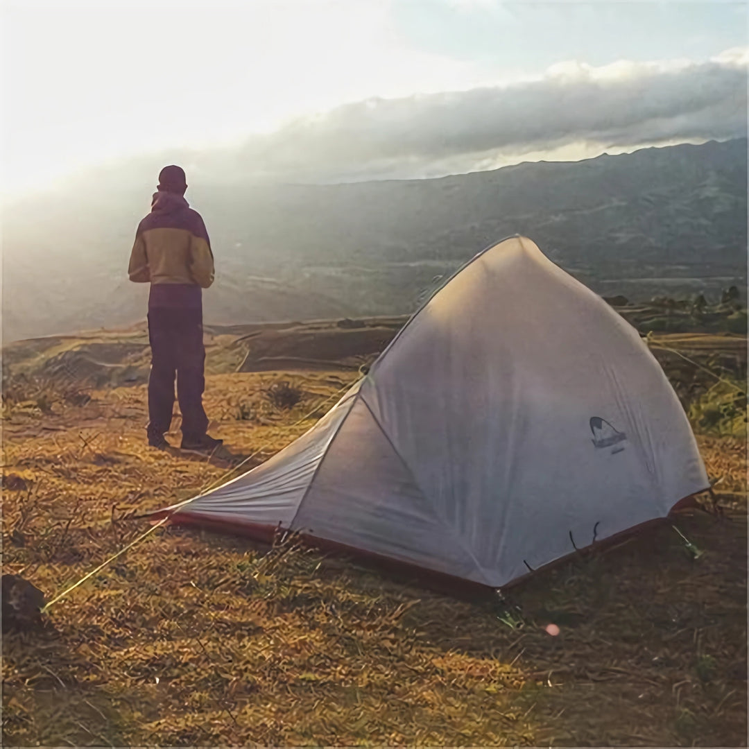 A person stands on a hilltop overlooking a mountain range and valley. In the picture, we can see a tent, which is set up on an open ground. The person stands next to the tent, seemingly admiring the magnificent view.