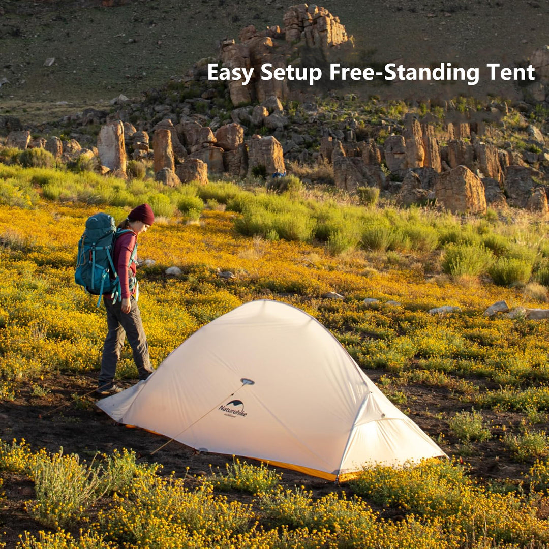 A man sets up a white tent with the "Naturehike" brand logo on a meadow full of yellow wildflowers. The background is a rocky terrain, and under the sunlight, the whole scene looks peaceful and natural.