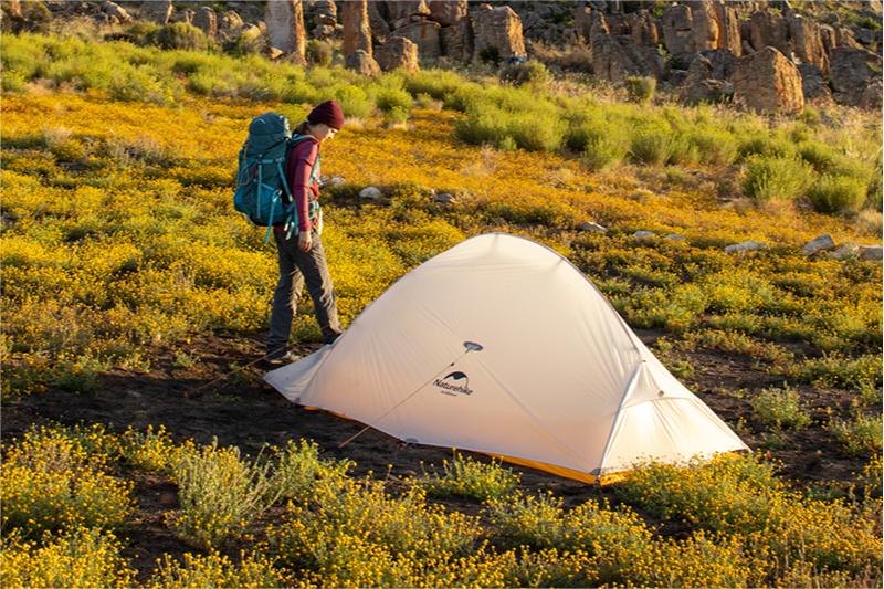 A woman sets up a white tent with the "Naturehike" brand logo on a meadow full of yellow wildflowers. The background is a rocky terrain, and the whole scene looks peaceful and natural under the sunlight.