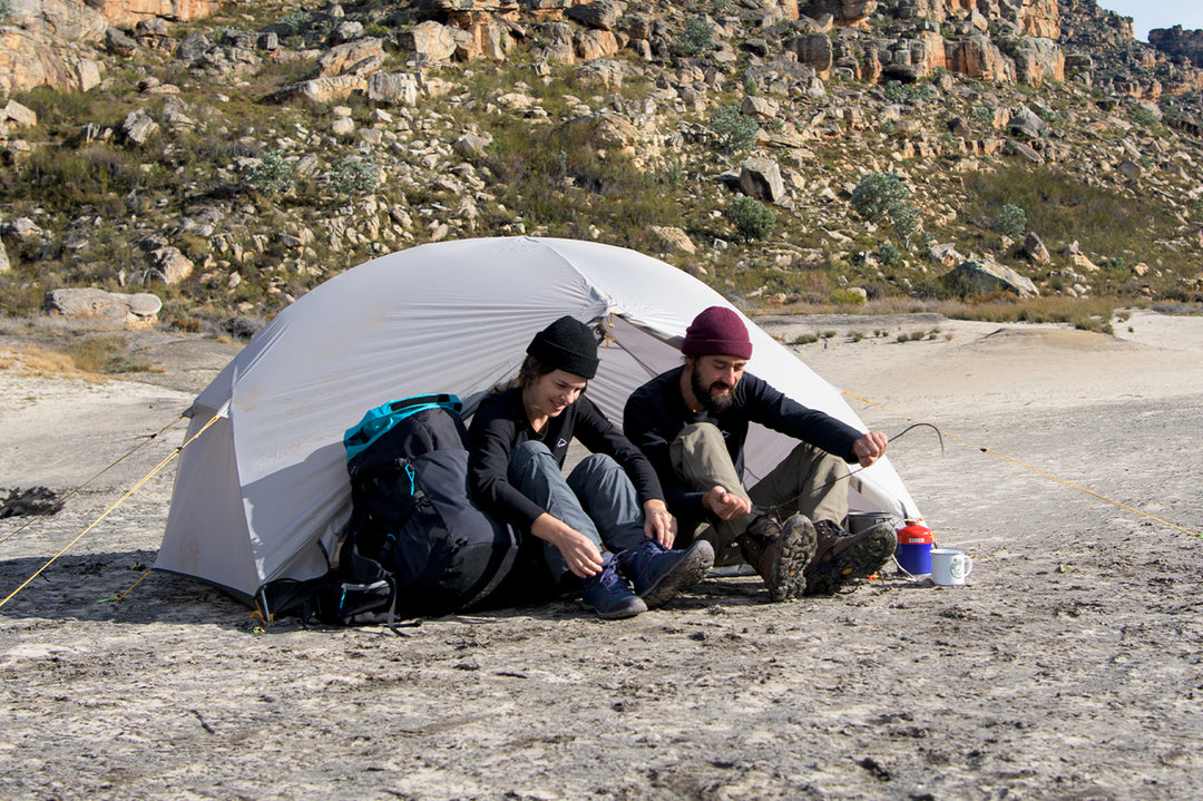 A pair of friends are sitting in front of the tent, they are enjoying the fun of camping. Surrounded by some rocks and vegetation, it gives a sense of nature and tranquility.