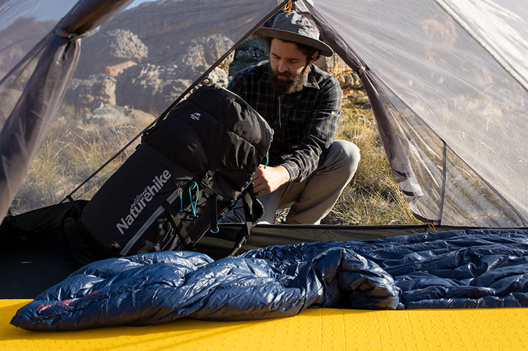 A man is sitting in a tent, packing his backpack. There are some items inside the tent, such as sleeping bags and backpacks. The whole scene gives people a feeling of comfort and relaxation.